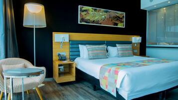 Habitación de hotel con cama, mesita de noche, lámpara, sillón y una obra de arte en la pared.