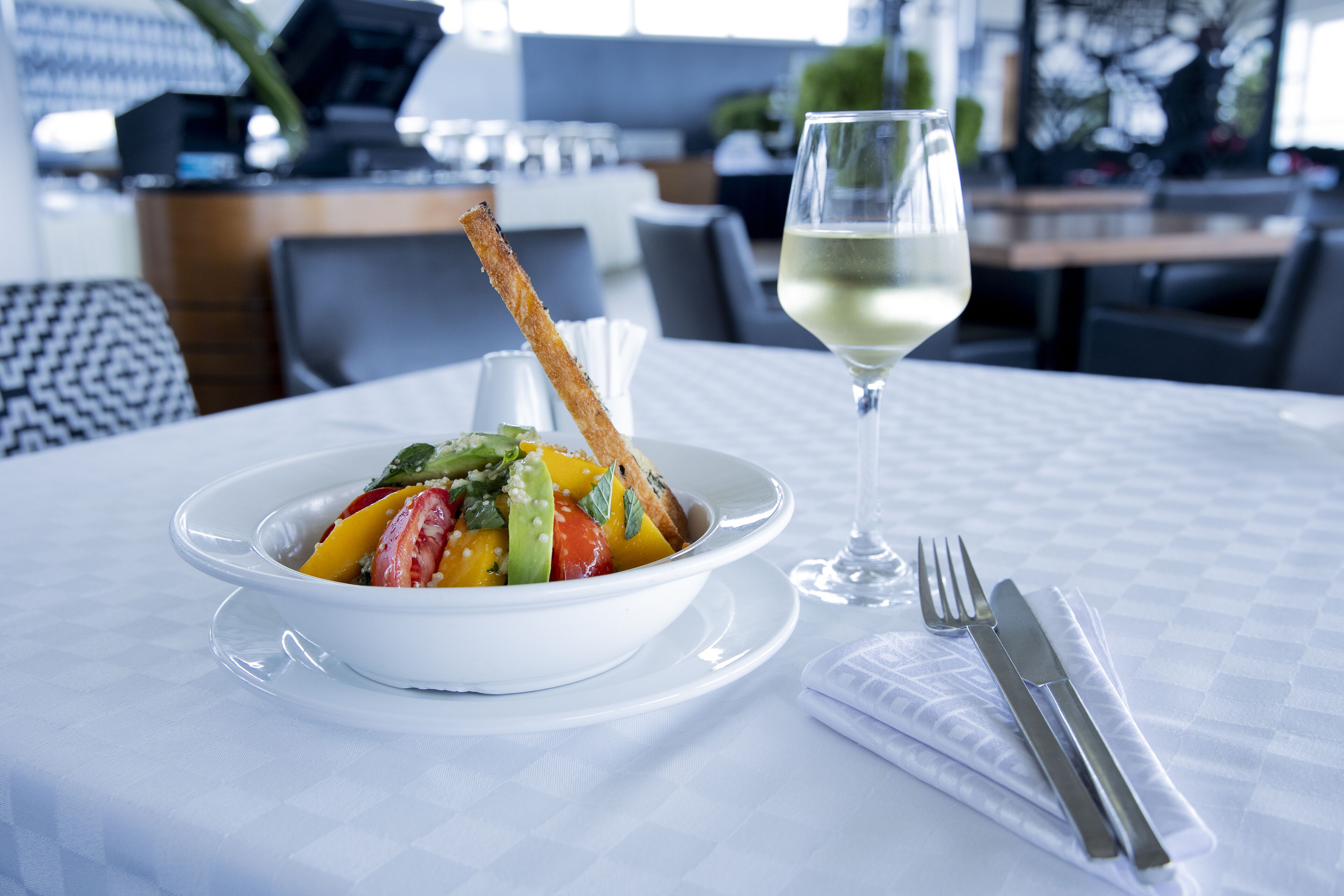 A dining table with a white tablecloth, a bowl of fruit salad, a glass of white wine, and cutlery.
