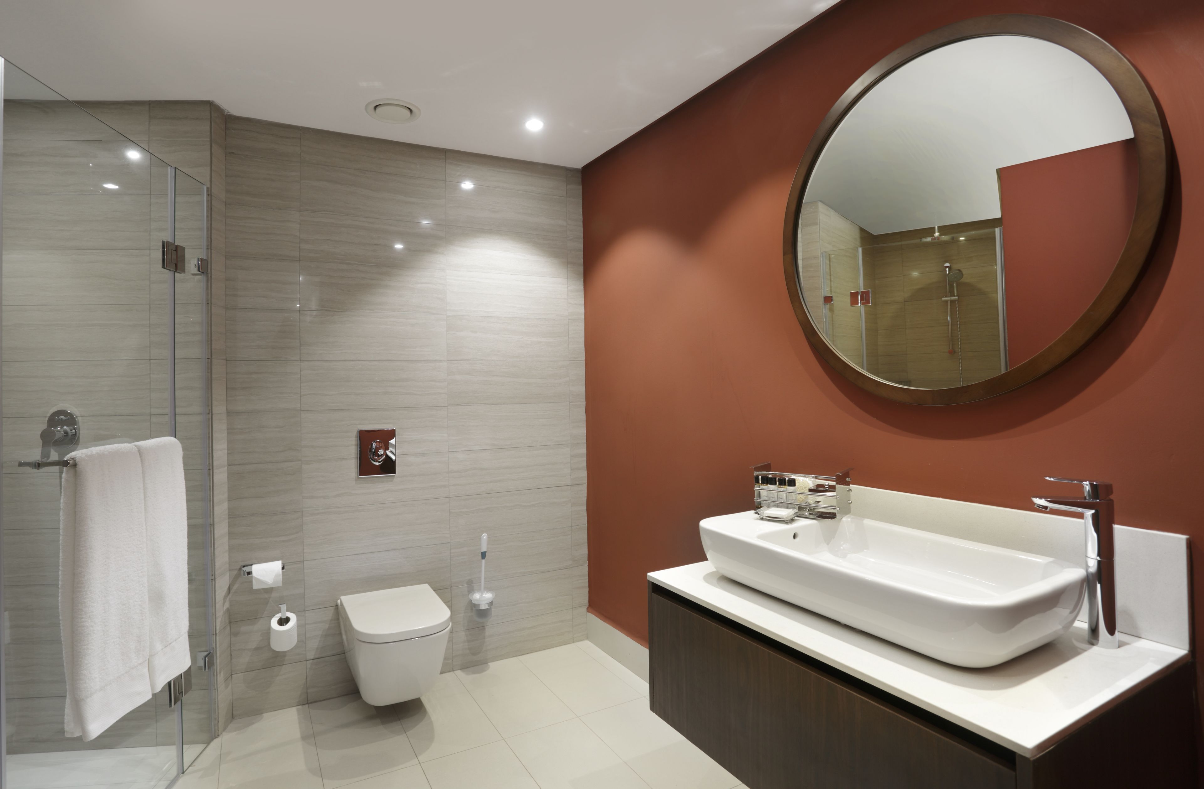 A modern bathroom with a glass shower enclosure, a white toilet, a large oval mirror, and a white sink with a dark cabinet.