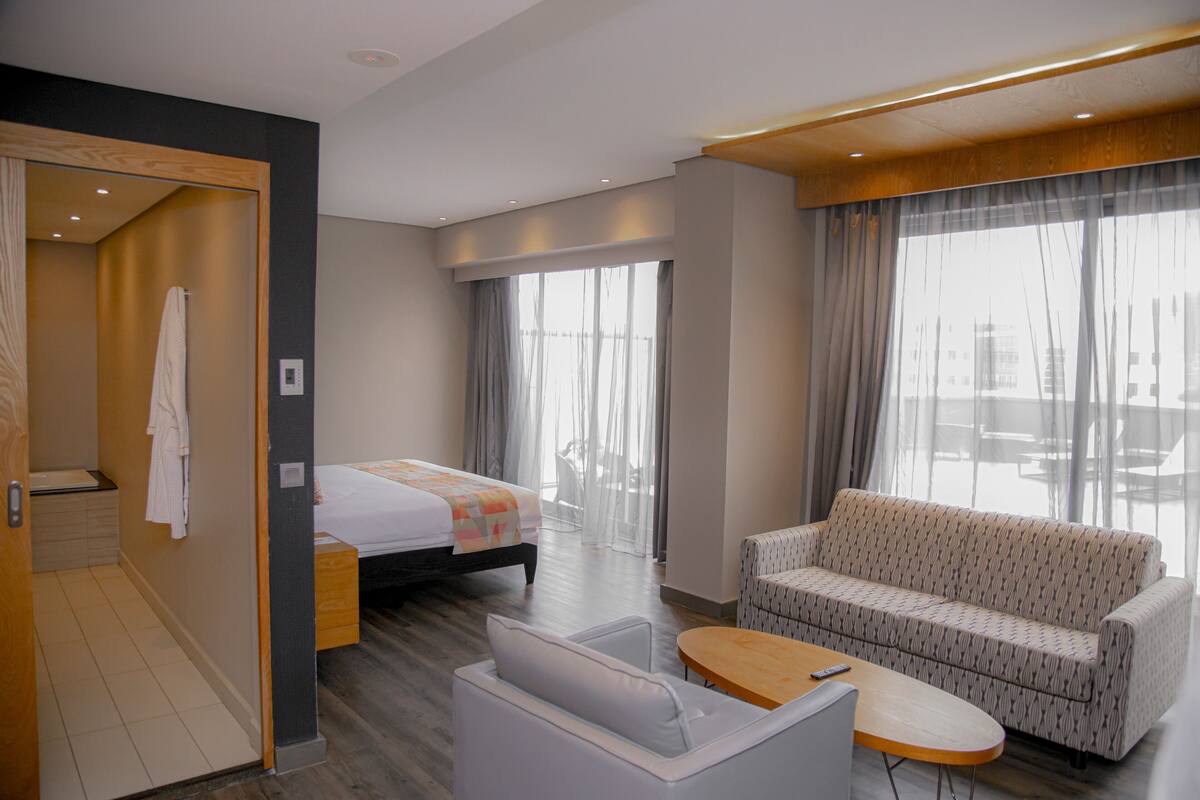 A hotel room with a bed, a sofa, a small table, and a view of the city through large windows.