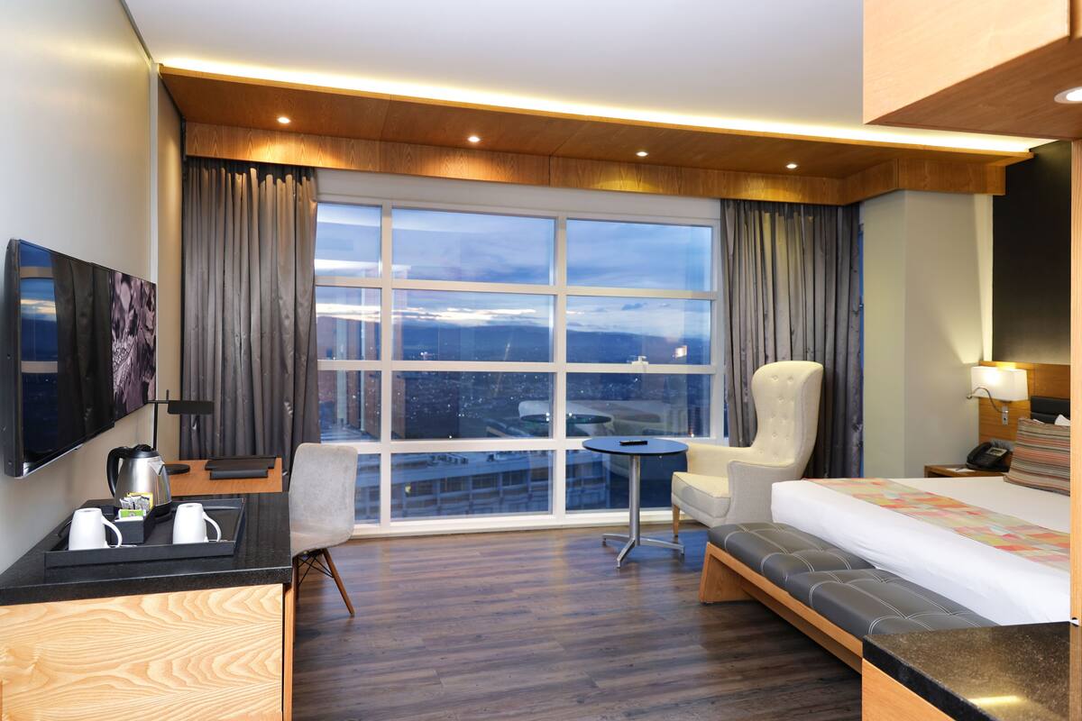 A modern hotel room with a large window, a bed, a desk with a kettle, a chair, and a small table.