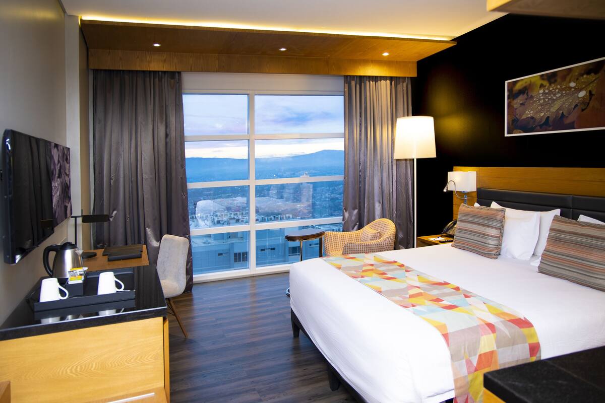 A hotel room with a large bed, a desk with a coffee maker, a chair, and a view of a cityscape through a large window.