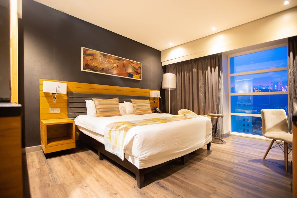 A hotel room with a large bed, a nightstand, a lamp, a chair, and a city view through the window.
