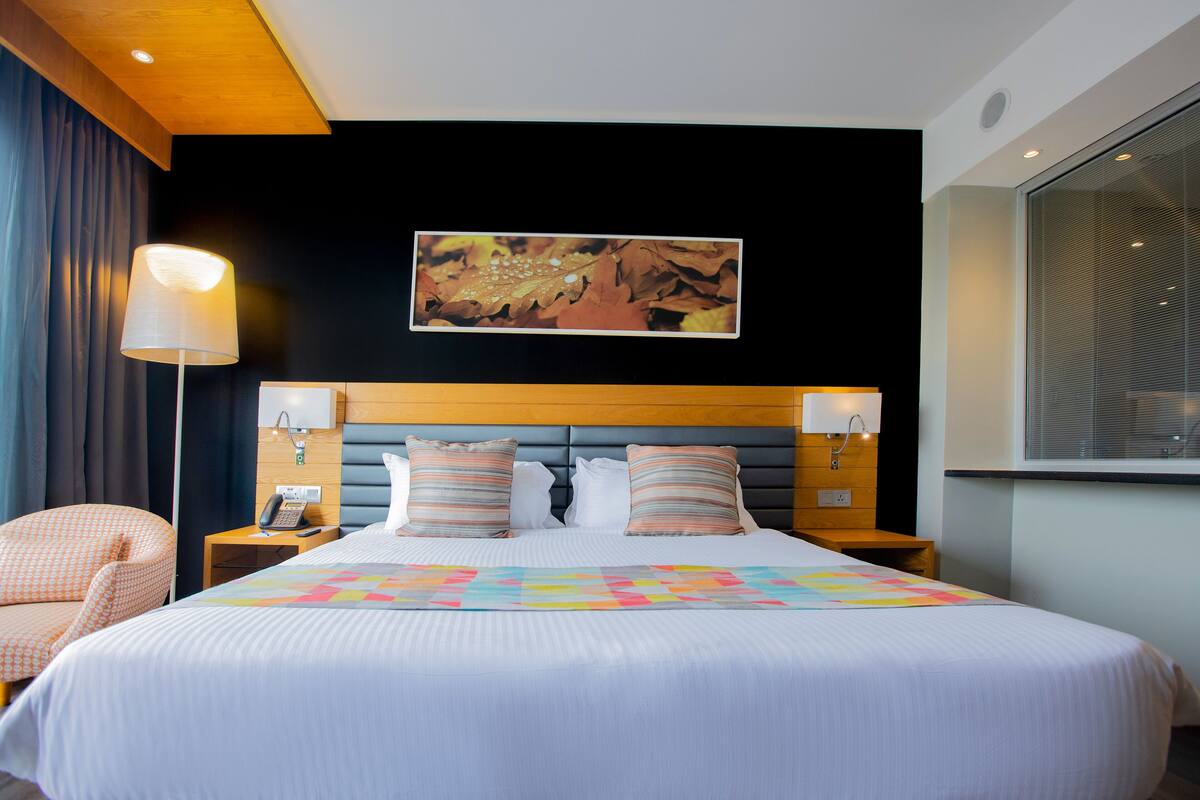 A hotel room with a large bed, a headboard with a wooden finish, a bedside table with a lamp, and a wall-mounted artwork.