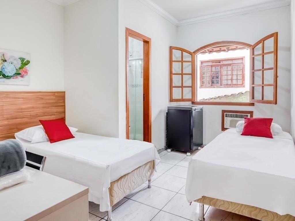 Quarto de hotel com duas camas de solteiro, uma mesinha, aquecedor e uma janela com persianas de madeira.