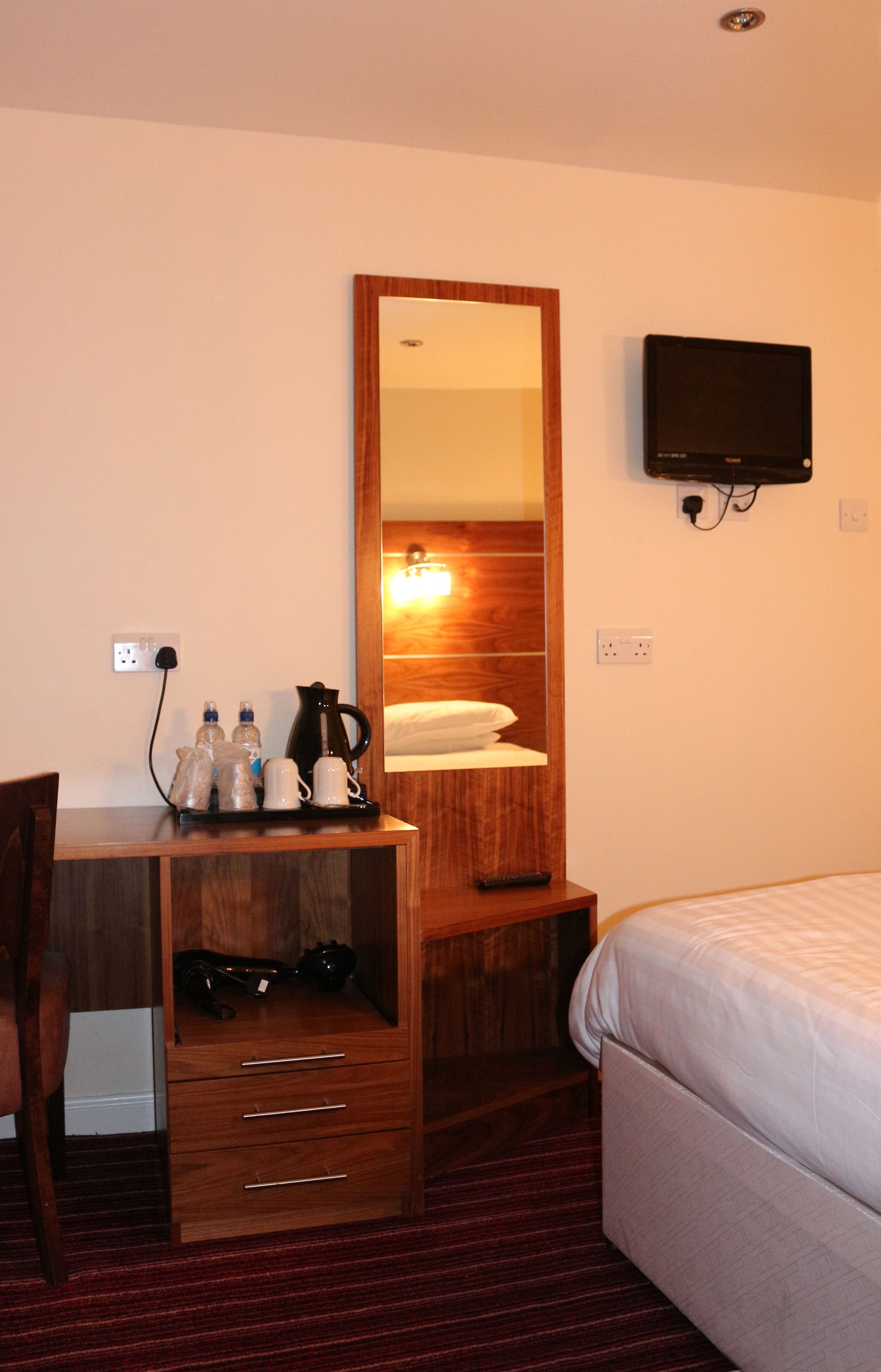A hotel room with a wooden desk, a mirror, a television, a bed, and a nightstand with a kettle and cups.