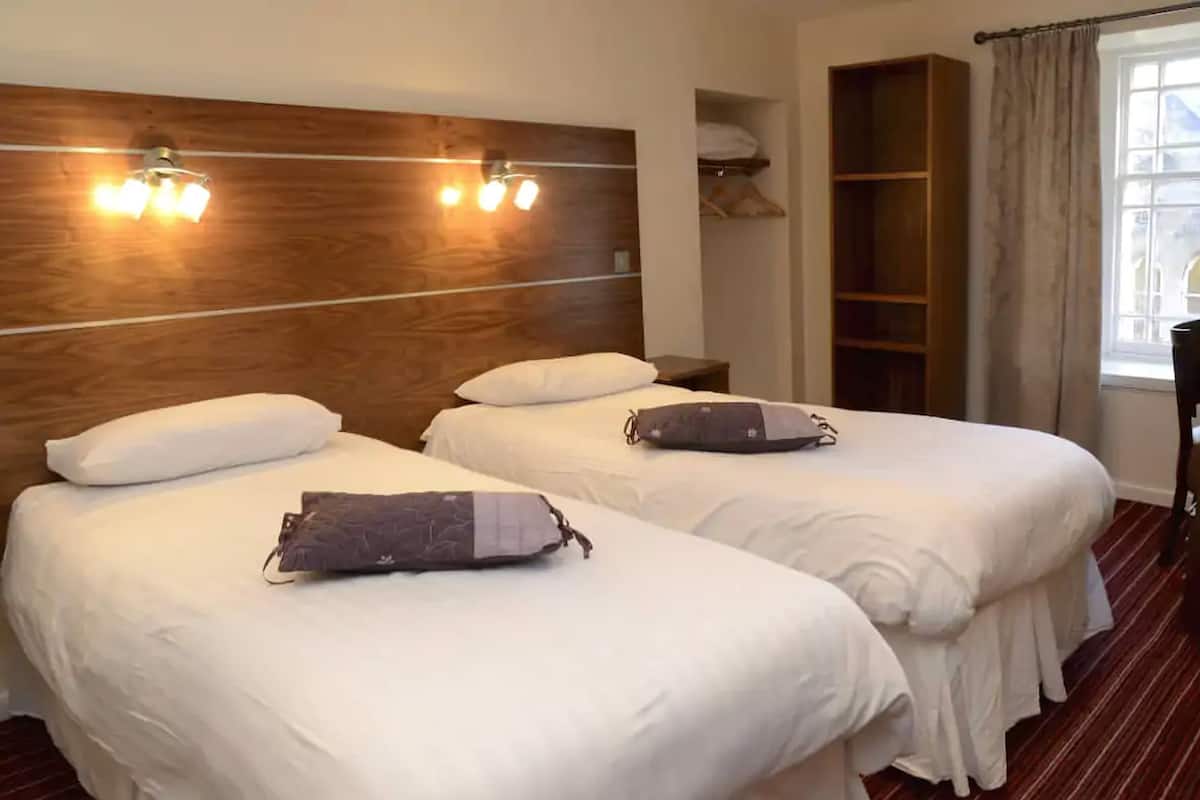 Ein Hotelzimmer mit zwei Betten, einem Holzkopfteil, Nachttischlampen und einem Fenster mit Vorhängen.