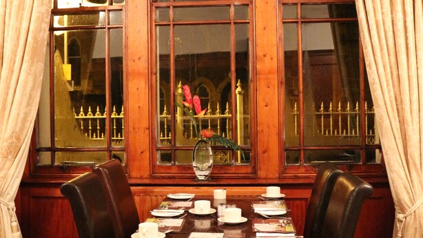 A dining area with a table set for four, two chairs, a window with curtains, and a view of a building with a fence. - County Hotel (Stornoway)