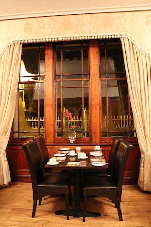 A dining area with a table set for four, two chairs, a window with curtains, and a view of a building with a fence. - County Hotel (Stornoway)