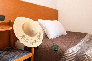 Una cama con colcha marrón, almohada blanca, cabecera de madera, mesita de noche de madera, silla azul y sombrero de paja con la inscripción 'Hello Goodnight'.