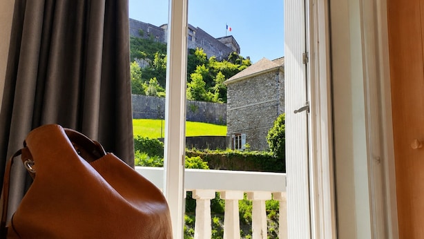 Ein Zimmer mit einem Bett, Blick auf ein Steinhaus und einer braunen Ledertasche auf dem Bett.