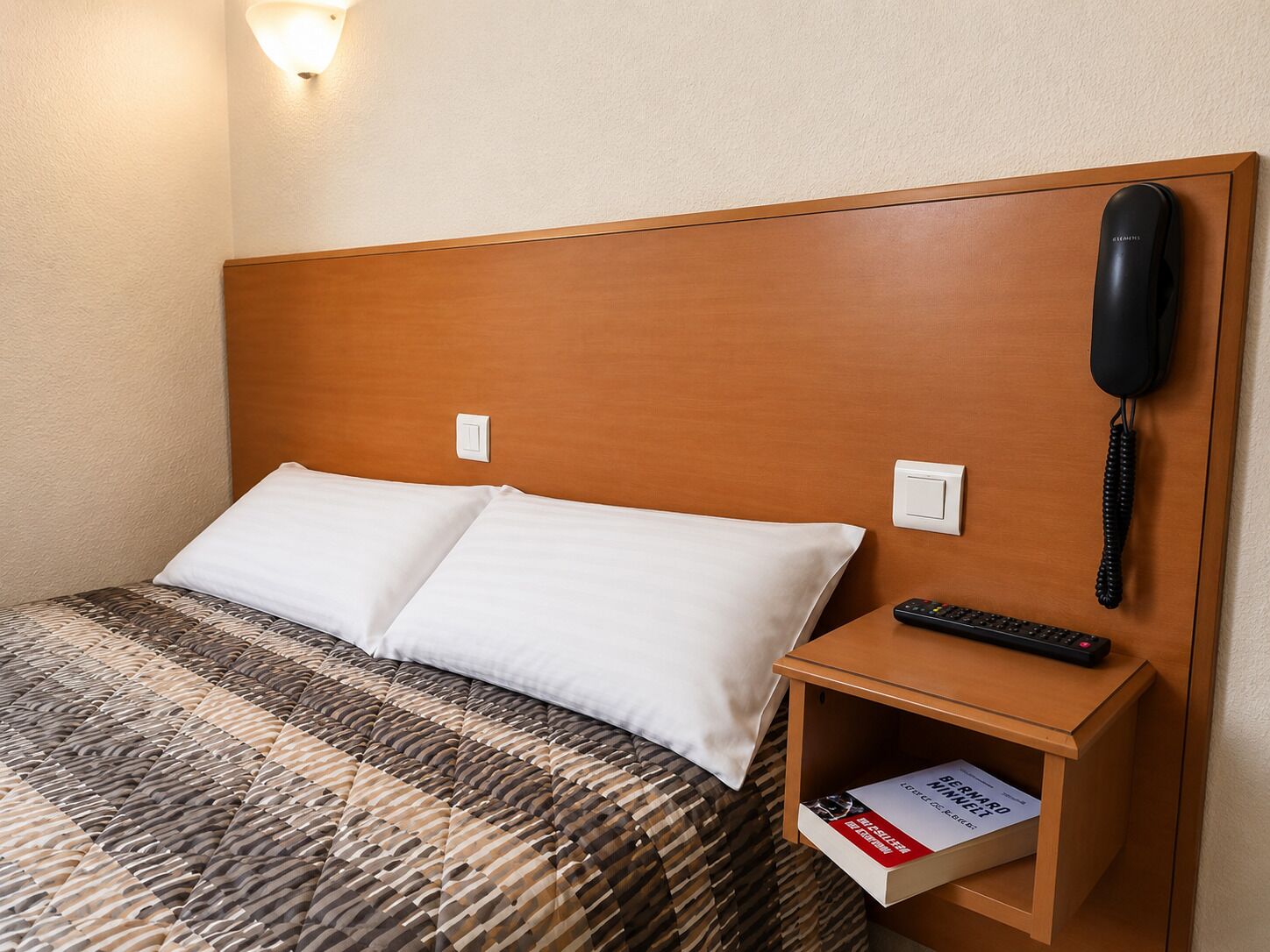 Une chambre d’hôtel avec une tête de lit en bois, un lit avec des oreillers blancs, une table de chevet avec une télécommande et un téléphone.