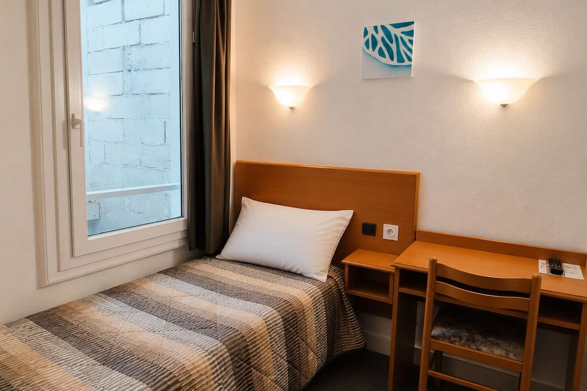 A single bed with a striped bedspread, a wooden headboard, a nightstand, a desk with a chair, and a wall-mounted light fixture.