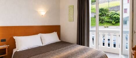 A hotel room with a large bed, a nightstand, a wall-mounted lamp, and a sliding glass door leading to a balcony with a view of greenery and a parking area.