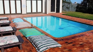 Une piscine rectangulaire bordée de carreaux, entourée d’une terrasse en briques. On y trouve des chaises longues avec des coussins et un oreiller à rayures.