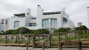A modern white building with multiple balconies and large windows, situated on a sandy beach with a wooden fence in the foreground. - On the Beach 1, Langebaan 2-Sleeper (Langebaan)