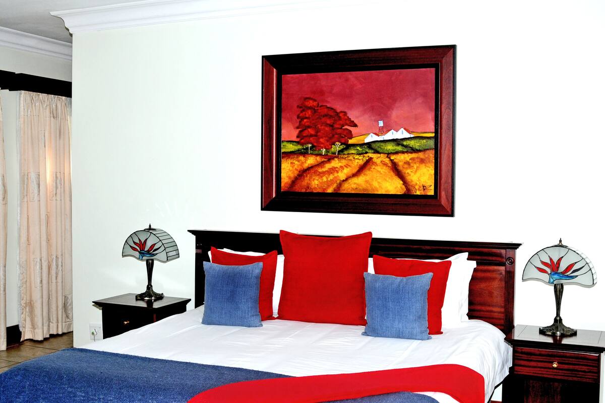 A bedroom with a bed, red and blue pillows, a painting, and two nightstands with lamps.
