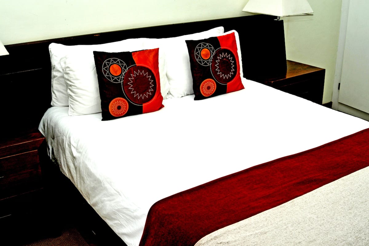 A neatly made bed with white and red bedding, two decorative pillows with geometric patterns, and a bedside table with a lamp.