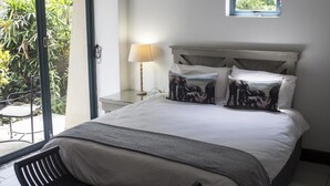 A bedroom with a bed, a nightstand with a lamp, a window with a view of greenery, and a small window above the bed. - Avalone Guest House (St. Lucia)