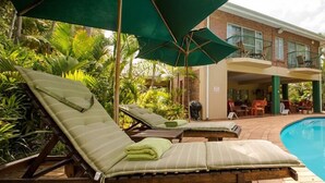 A poolside area with lounge chairs and umbrellas, a brick building with a balcony, and lush tropical vegetation. - Avalone Guest House (St. Lucia)