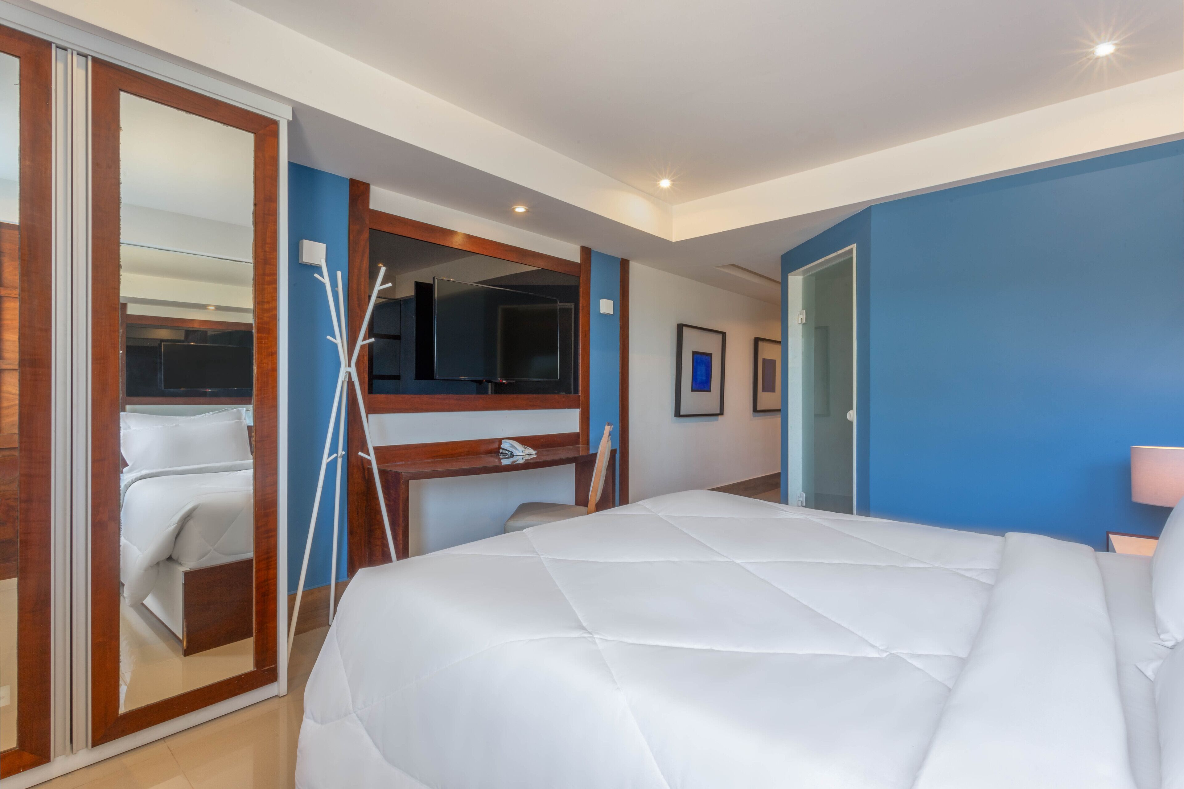 A hotel room with a large bed, a desk with a chair, a mirror, and a wardrobe.