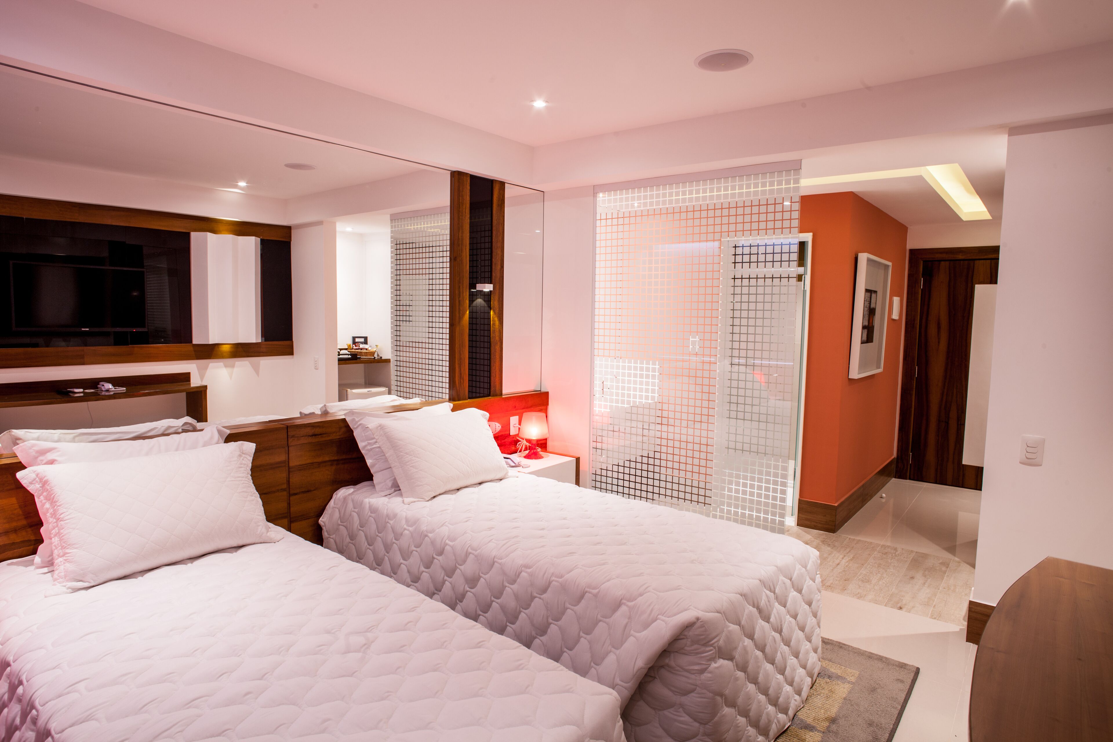 Una habitación de hotel moderna con dos camas, un espejo grande, un televisor y un baño con mampara de vidrio.