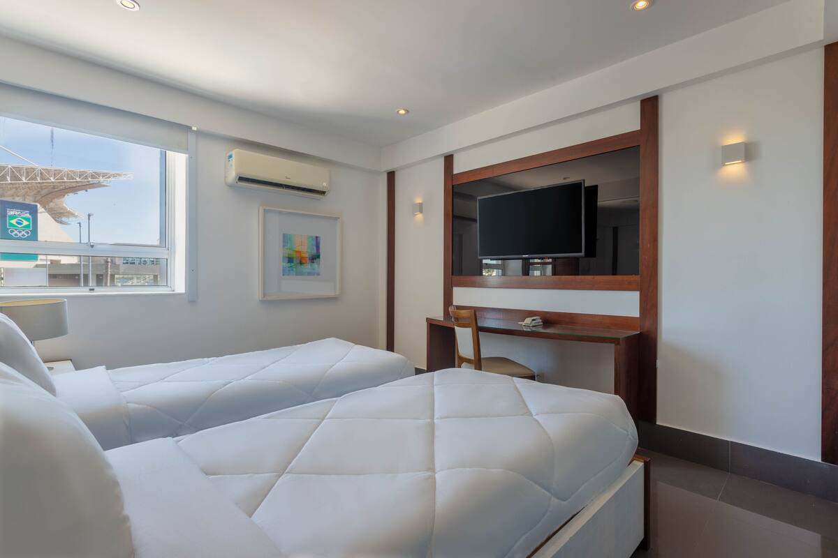 A hotel room with two beds, a TV, a desk, and a window with an air conditioning unit.
