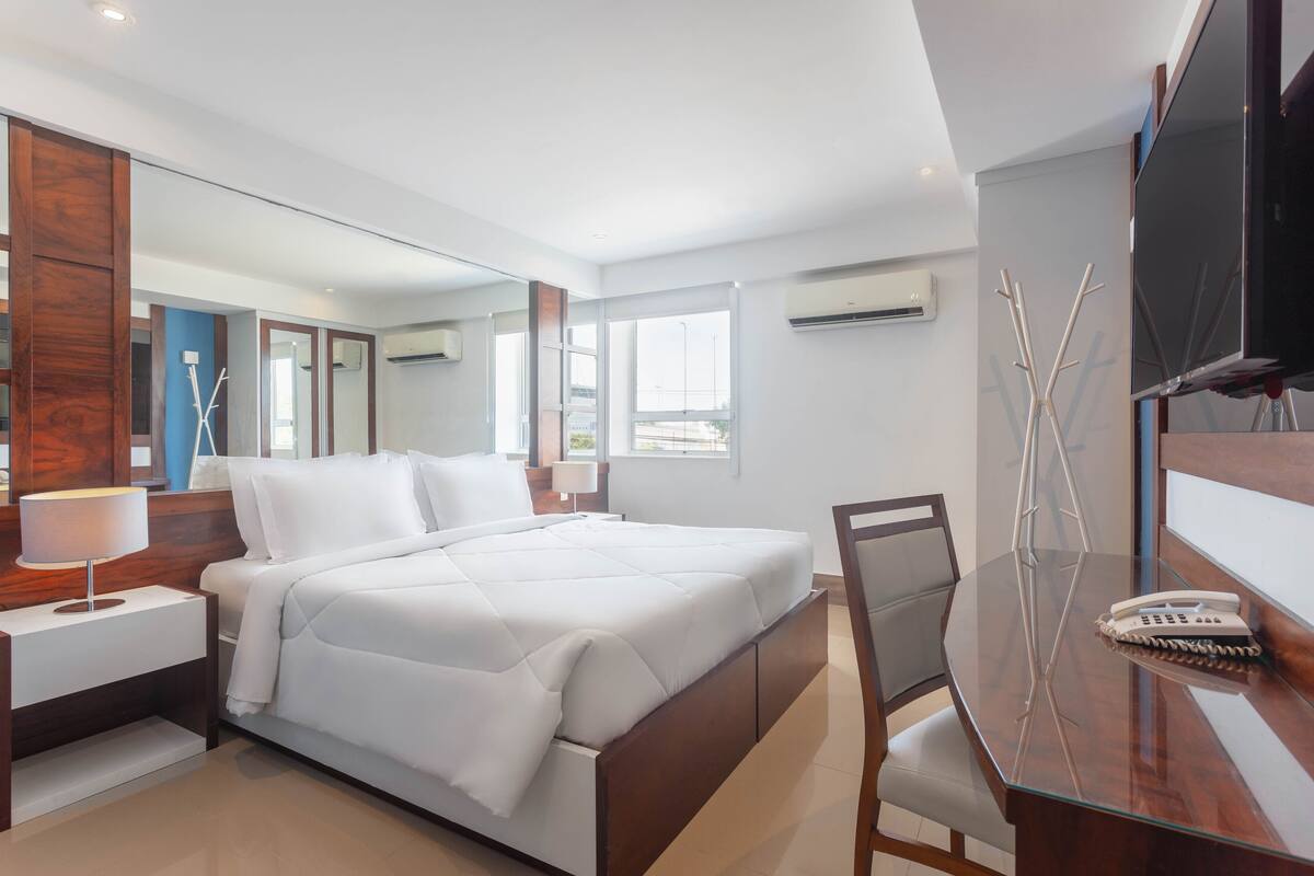 A modern hotel room with a large bed, a desk with a chair, a TV, and a large mirror.