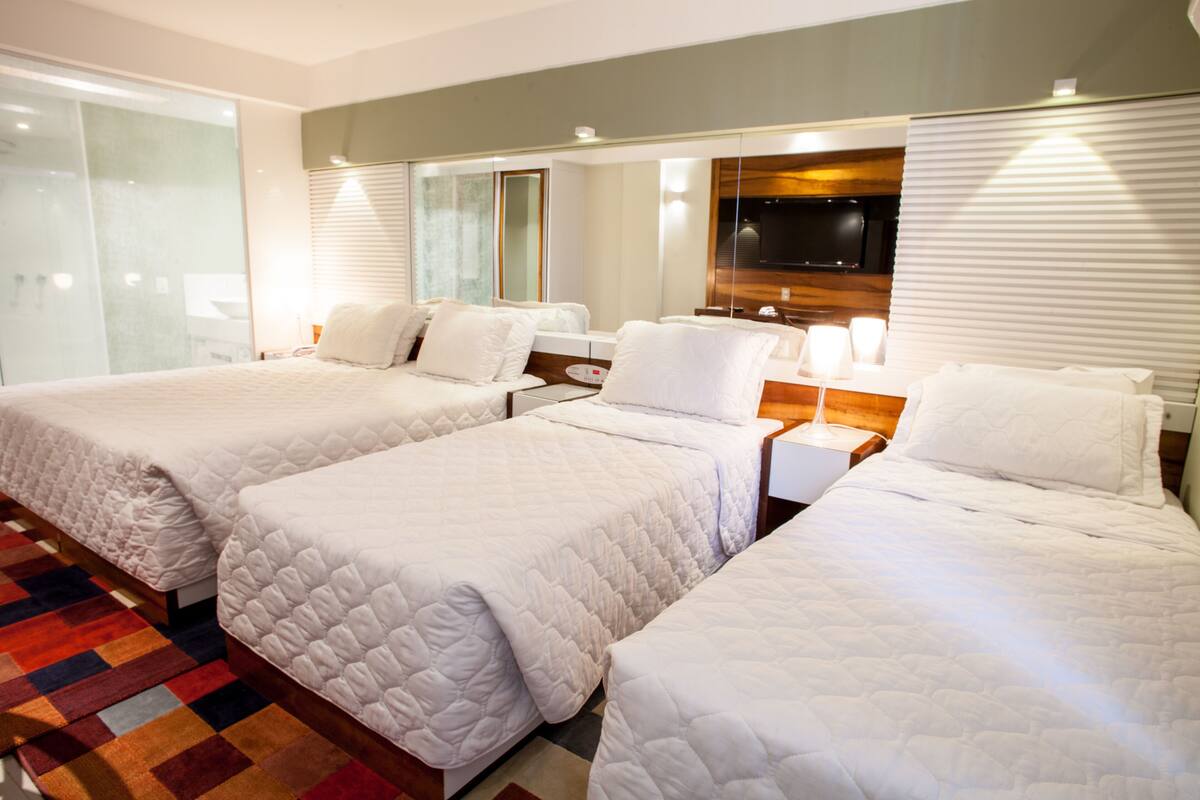 A hotel room with three beds, a TV, and a large window with blinds.