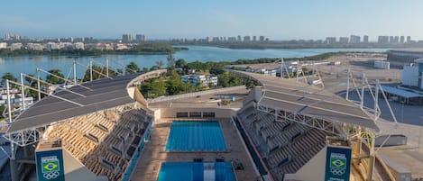 Un estadio olímpico de natación con una piscina grande, zonas de asientos y vistas a un horizonte urbano y una masa de agua.