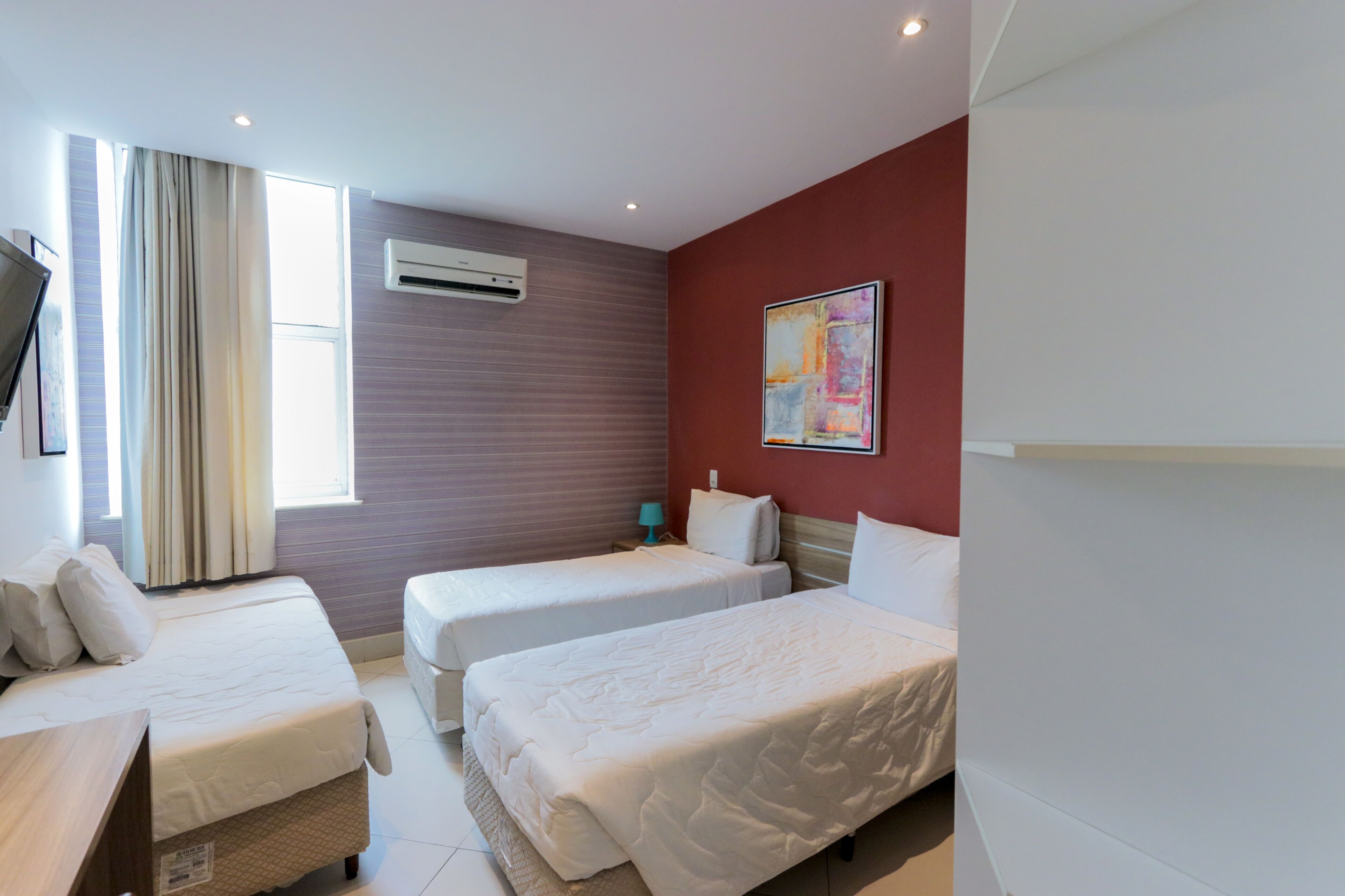 A hotel room with two single beds, a flat-screen TV, an air conditioning unit, and a window with curtains.