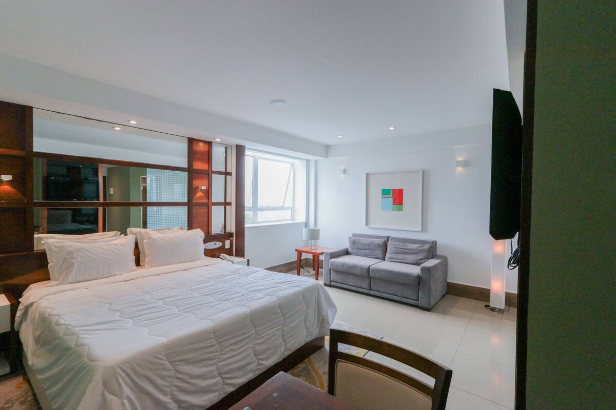 A modern hotel room with a large bed, a grey sofa, a small table, and a TV mounted on the wall.