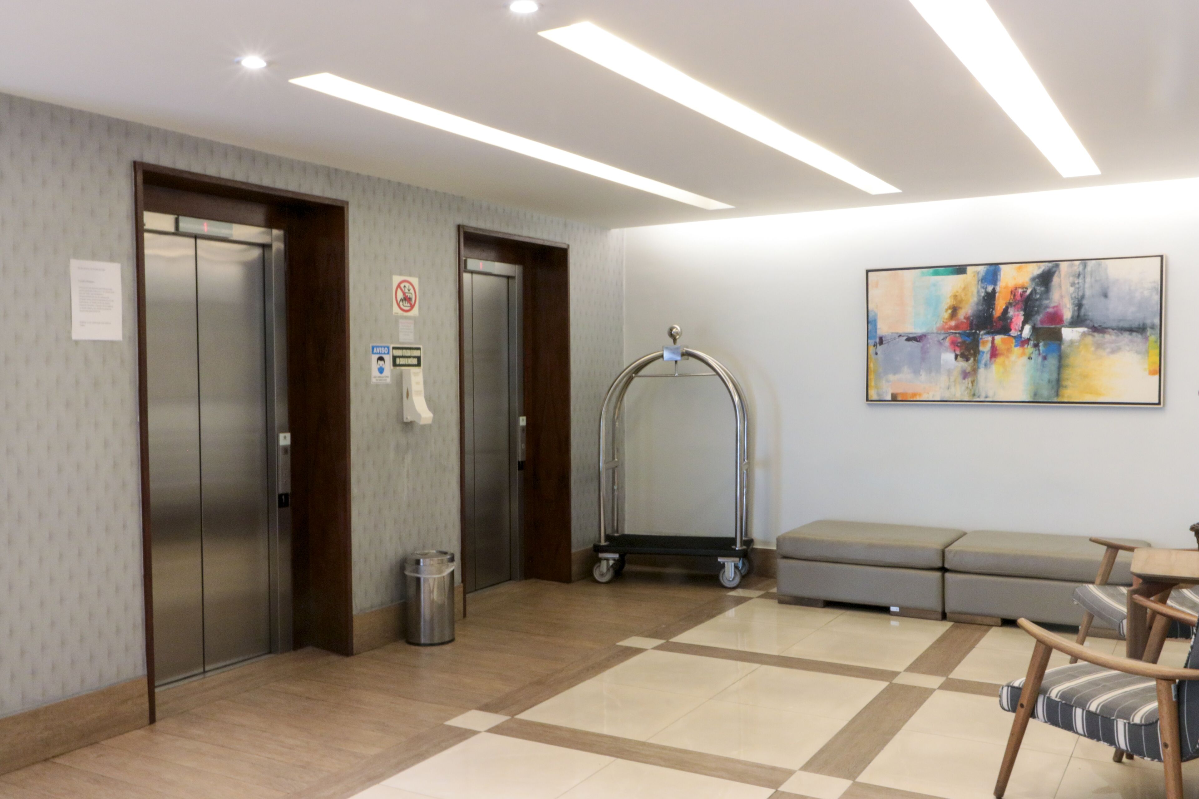A lobby with two elevators, a trash bin, a cart, a sofa, and a colorful abstract painting on the wall.