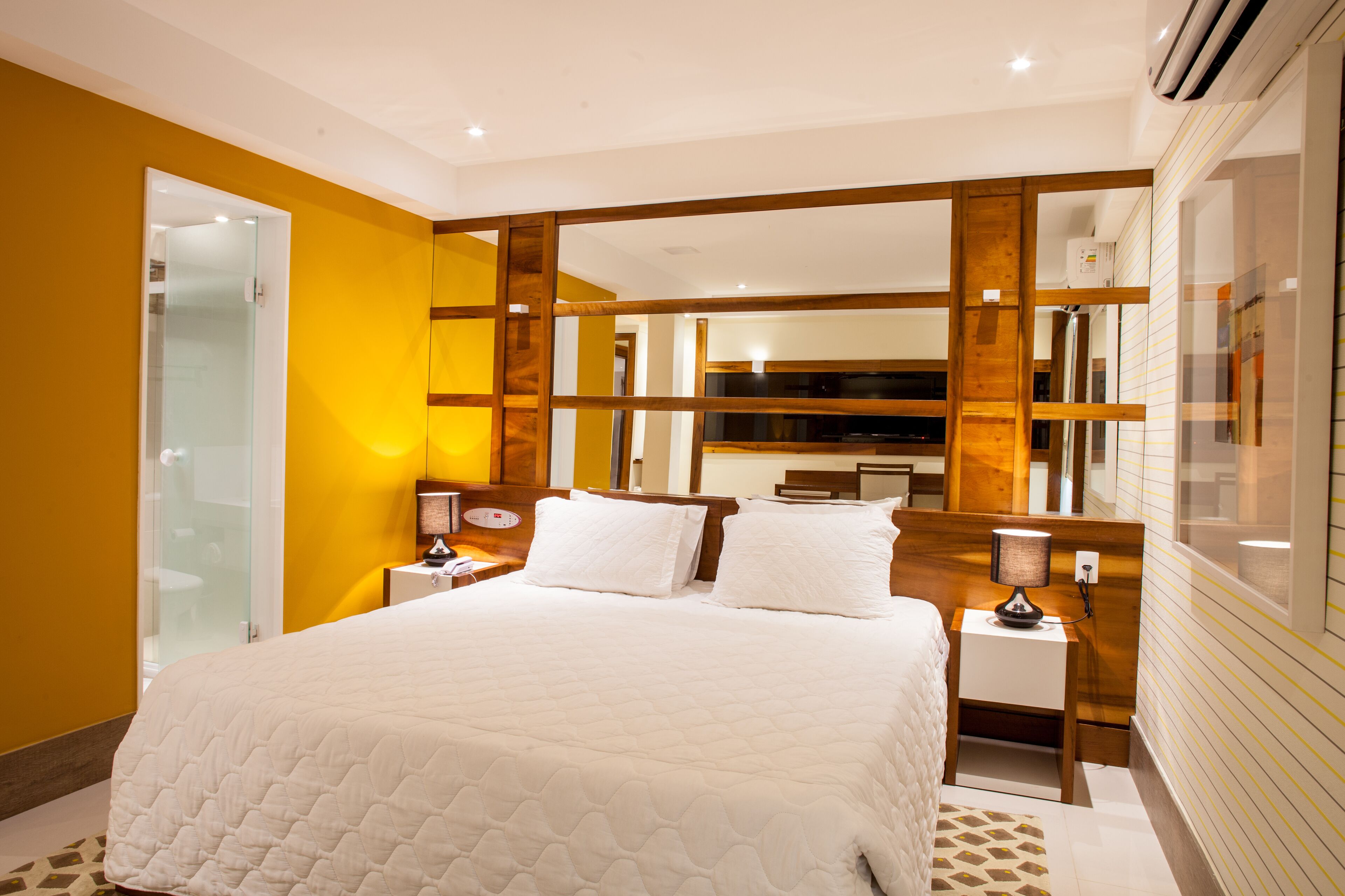 A modern hotel room with a large bed, wooden headboard, bedside lamps, and a glass shower enclosure.