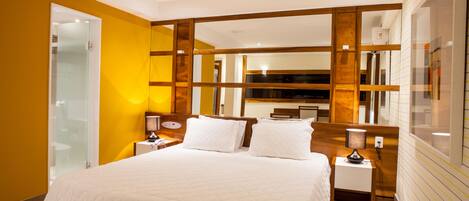 A modern hotel room with a large bed, wooden headboard, bedside lamps, and a glass shower enclosure.