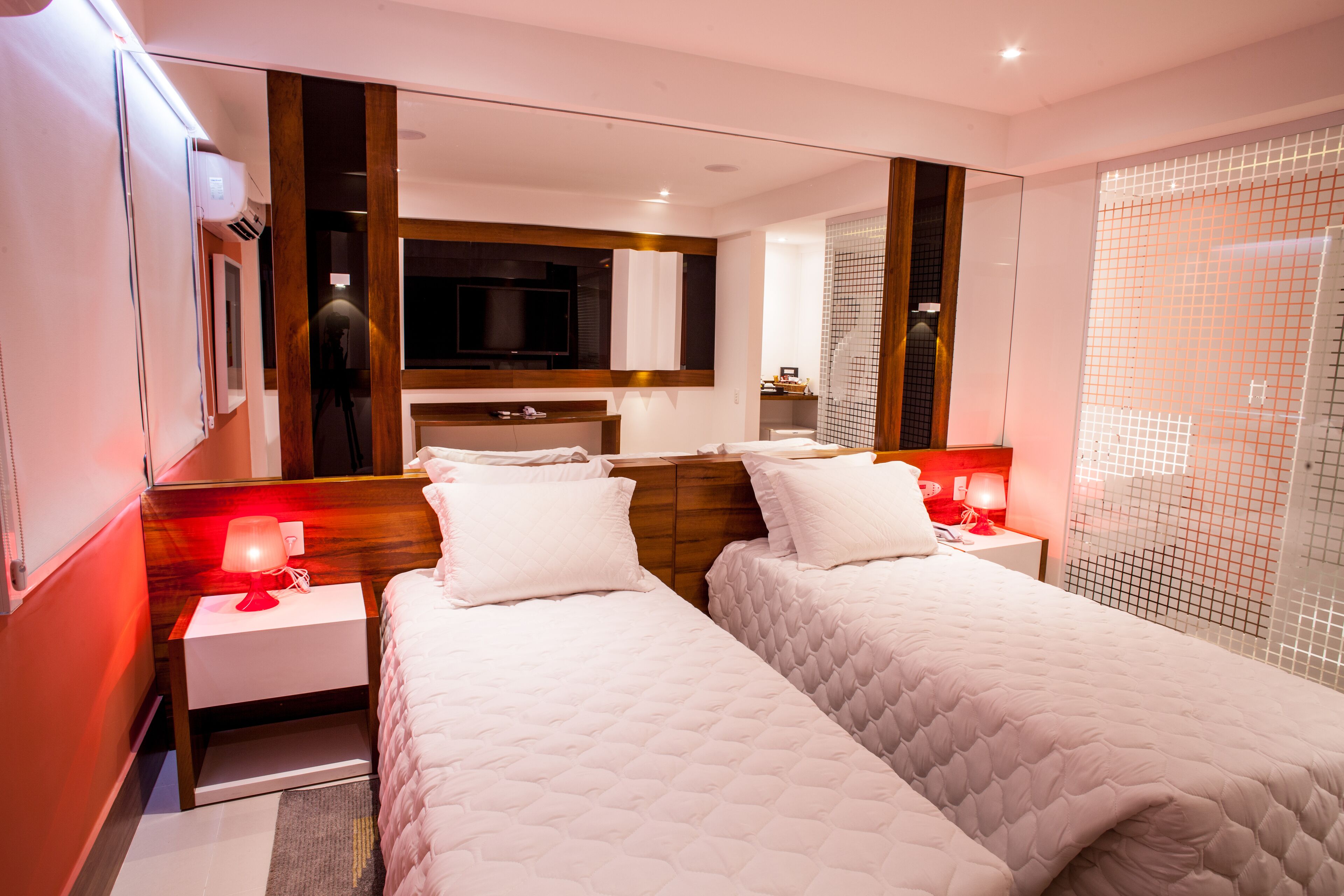 Quarto de hotel moderno com duas camas, televisão, espelho e chuveiro.