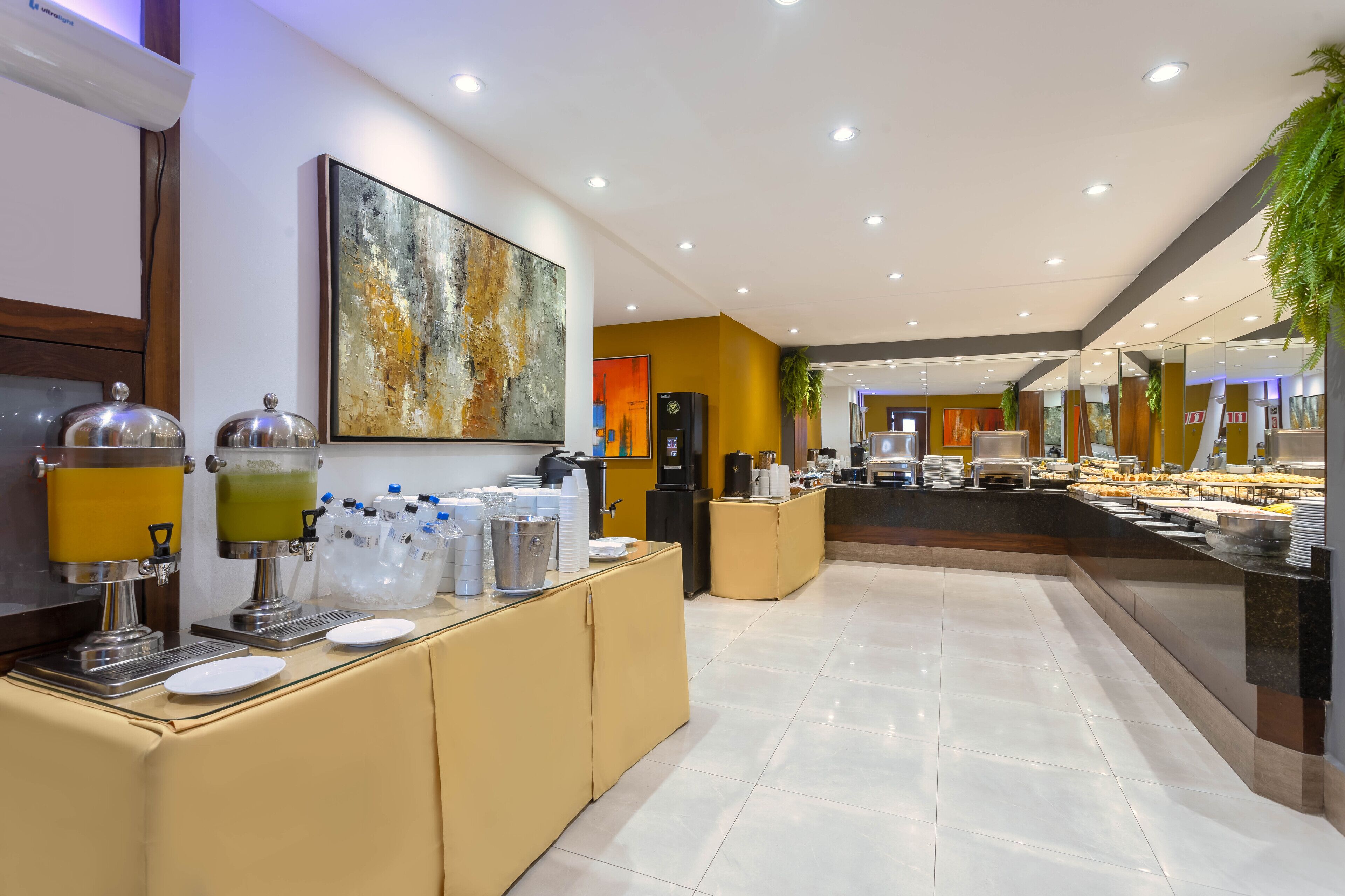 Un moderno vestíbulo de hotel con zona de buffet, incluyendo una estación de bebidas con dispensadores y un gran cuadro abstracto en la pared.