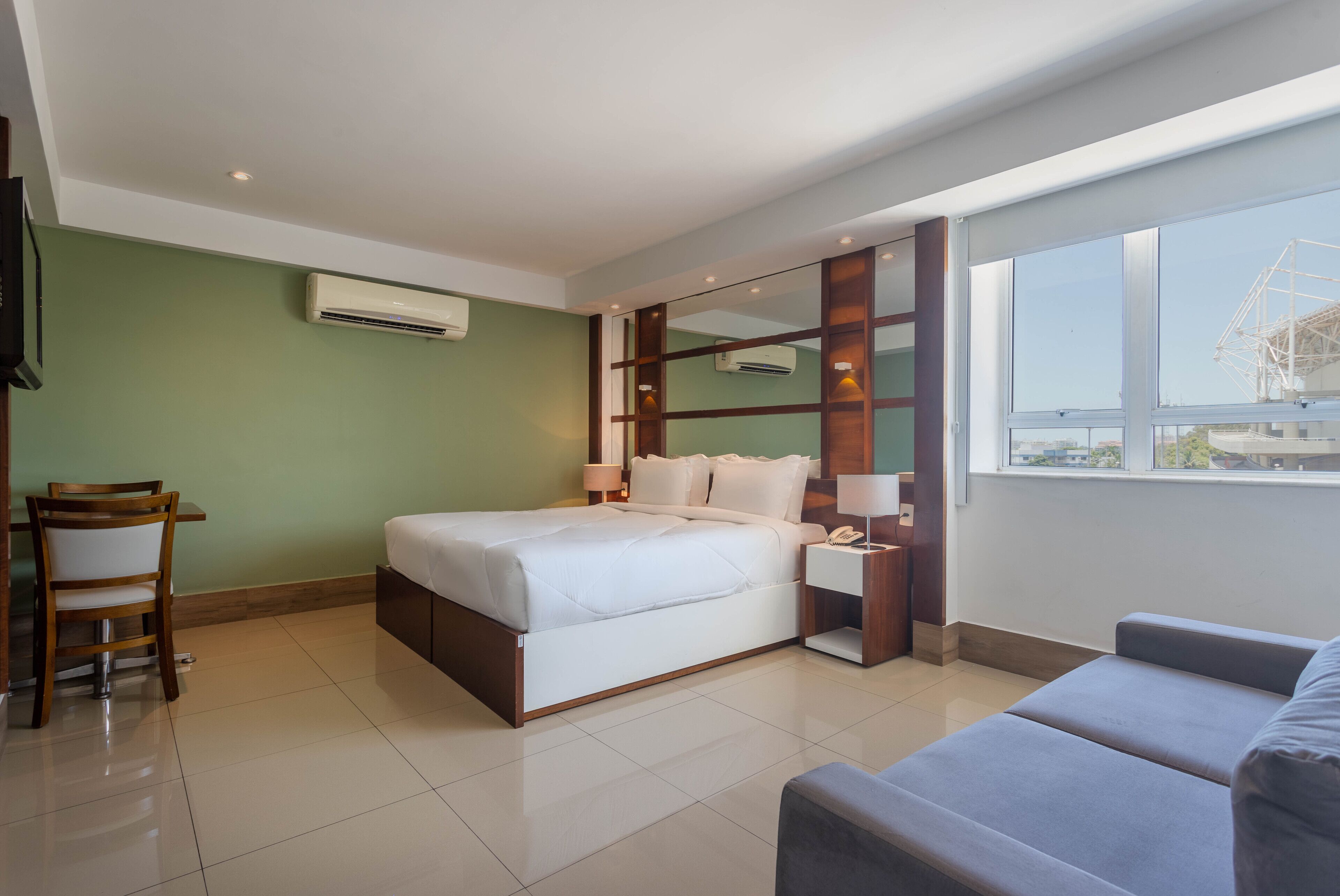 A modern hotel room with a large bed, a desk with a chair, a sofa, a large window, and an air conditioning unit.