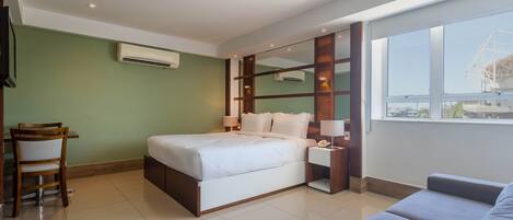 A modern hotel room with a large bed, a desk with a chair, a sofa, a large window, and an air conditioning unit.