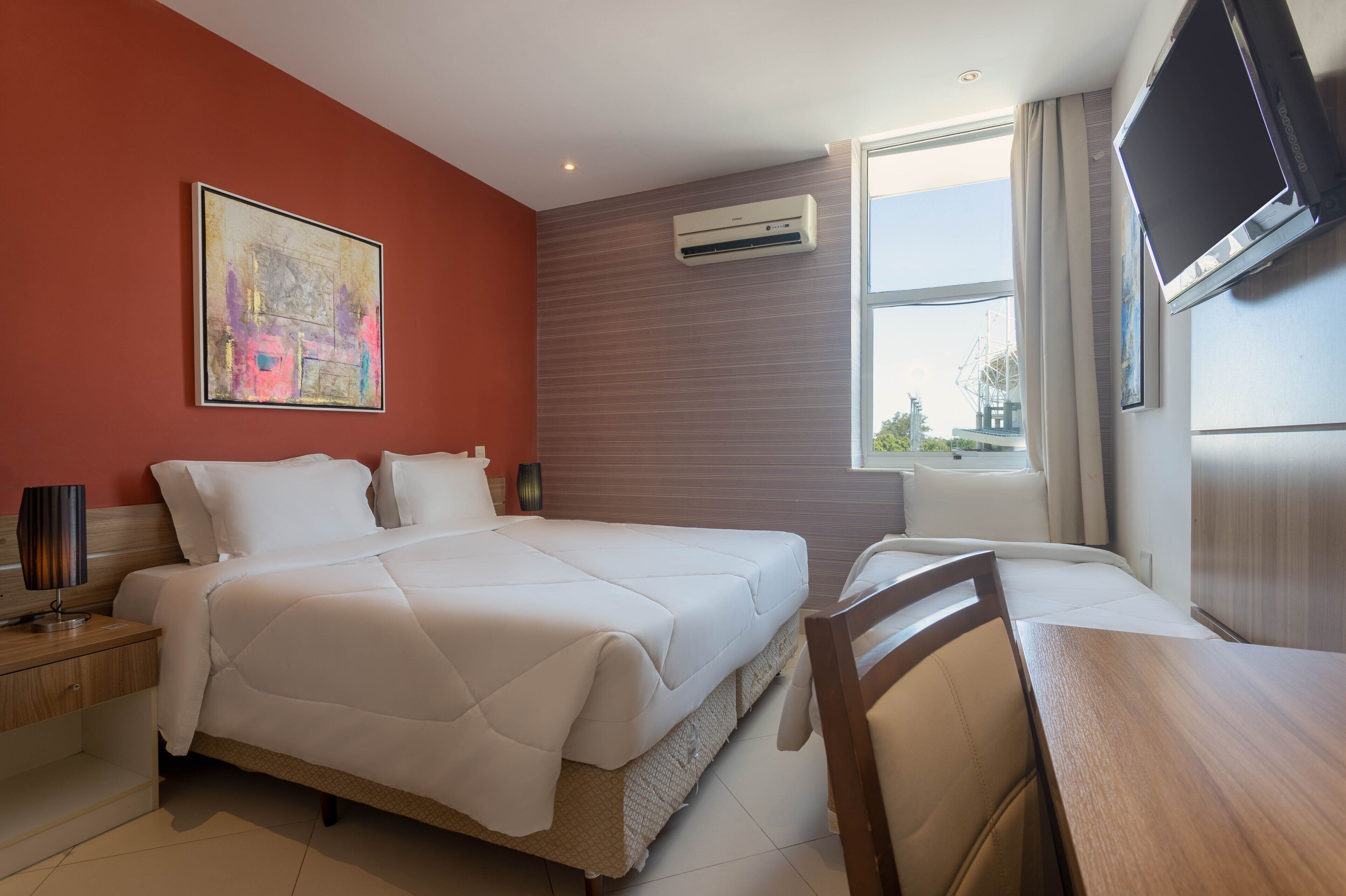 Habitación de hotel con cama, mesita de noche, lámpara, aire acondicionado, televisor y ventana con cortinas.