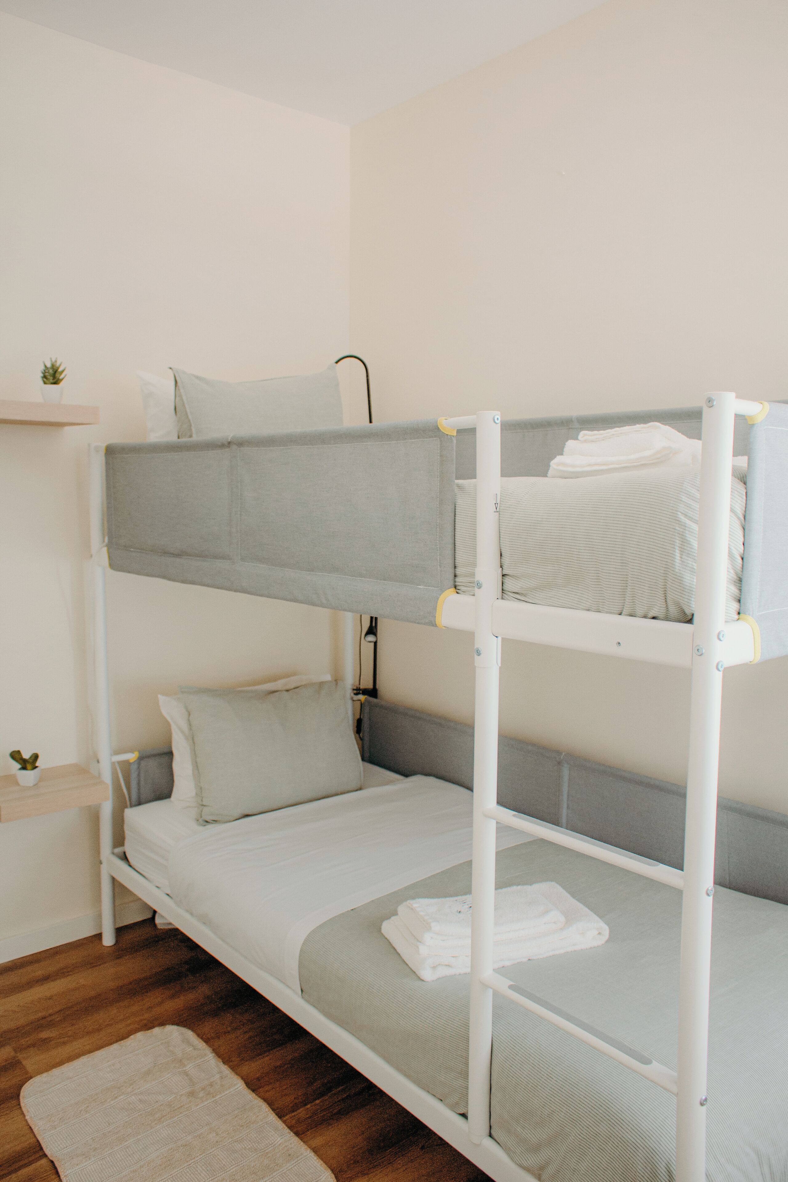 Un letto a castello con materasso grigio e biancheria bianca, un piccolo ripiano con una pianta e un pavimento in legno.