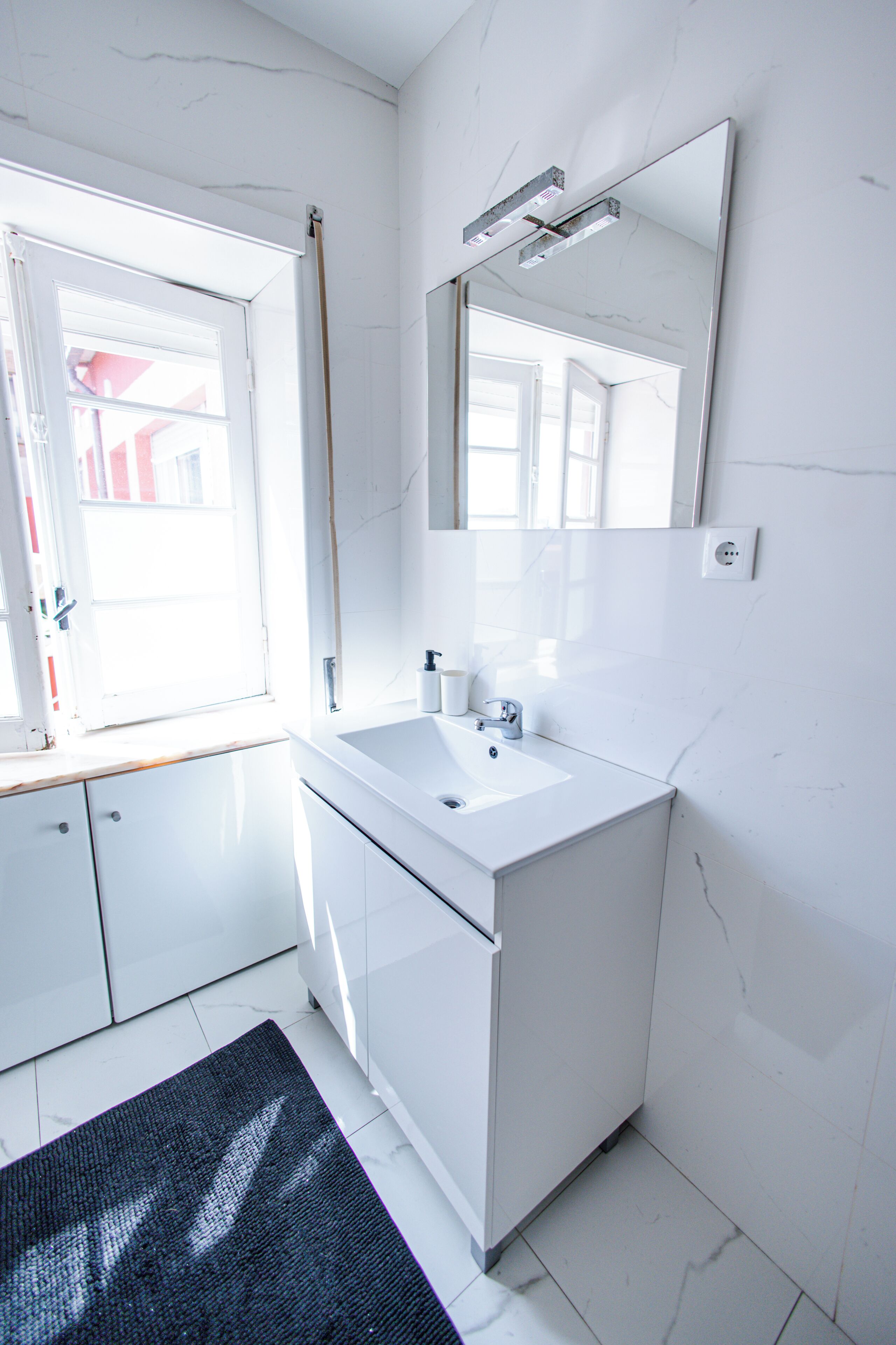 Un baño moderno con tocador blanco, un espejo grande y una ventana que deja entrar luz natural.