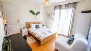 Un dormitorio con cama de madera, ropa de cama blanca, una mesita de noche con lámpara, una pequeña planta en maceta y una ventana con cortinas.