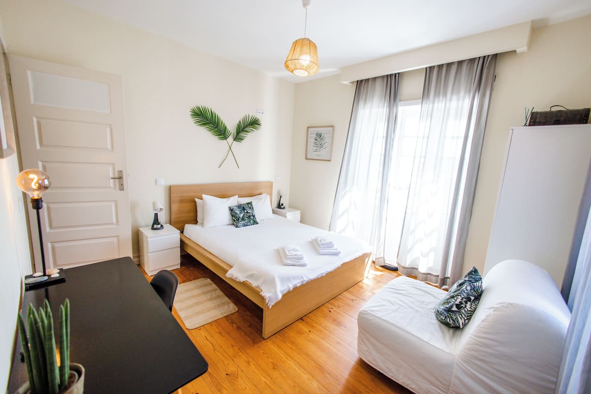 A bedroom with a wooden bed, white bedding, a bedside table with a lamp, a small potted plant, and a window with curtains.