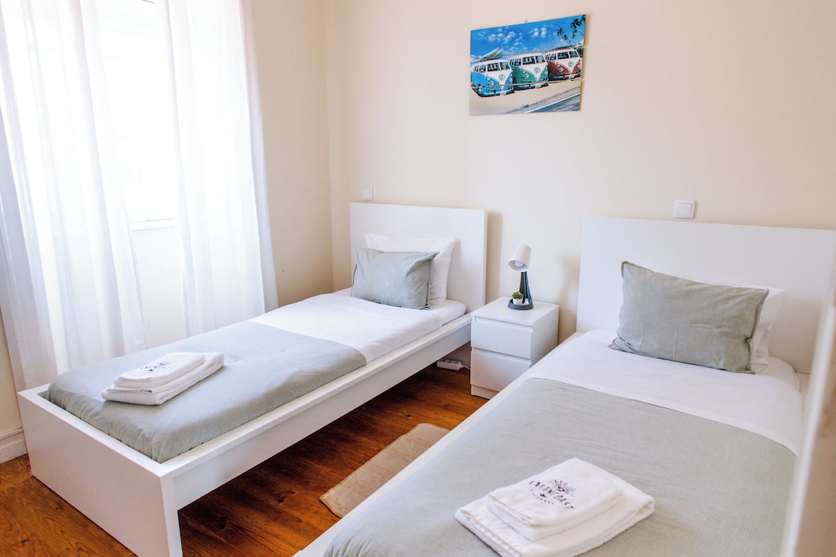A bedroom with two single beds, a bedside table with a lamp, a painting on the wall, and a window with curtains.