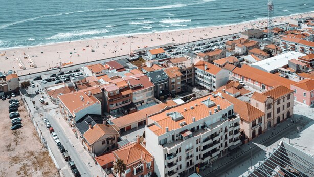 Vista aérea de un pueblo costero con una playa, edificios con techos de tejas rojas y un estacionamiento.