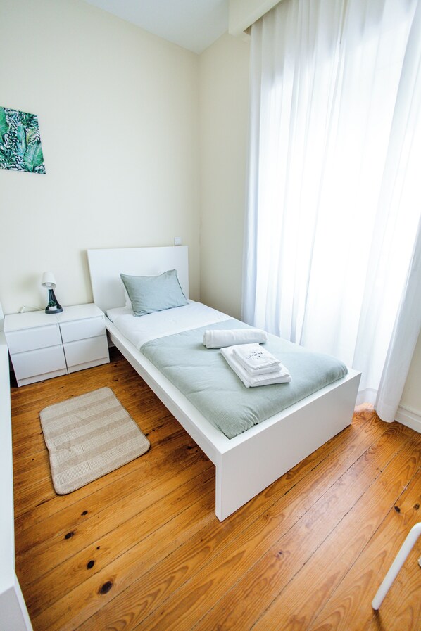 Un dormitorio minimalista con una cama individual, una mesita de noche y un ventanal con cortinas translúcidas.