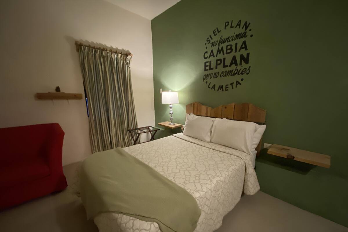Ein ordentlich bezogenes Bett mit einem gemusterten Überwurf, einer Nachttischlampe und einer Wand mit motivierenden Texten.