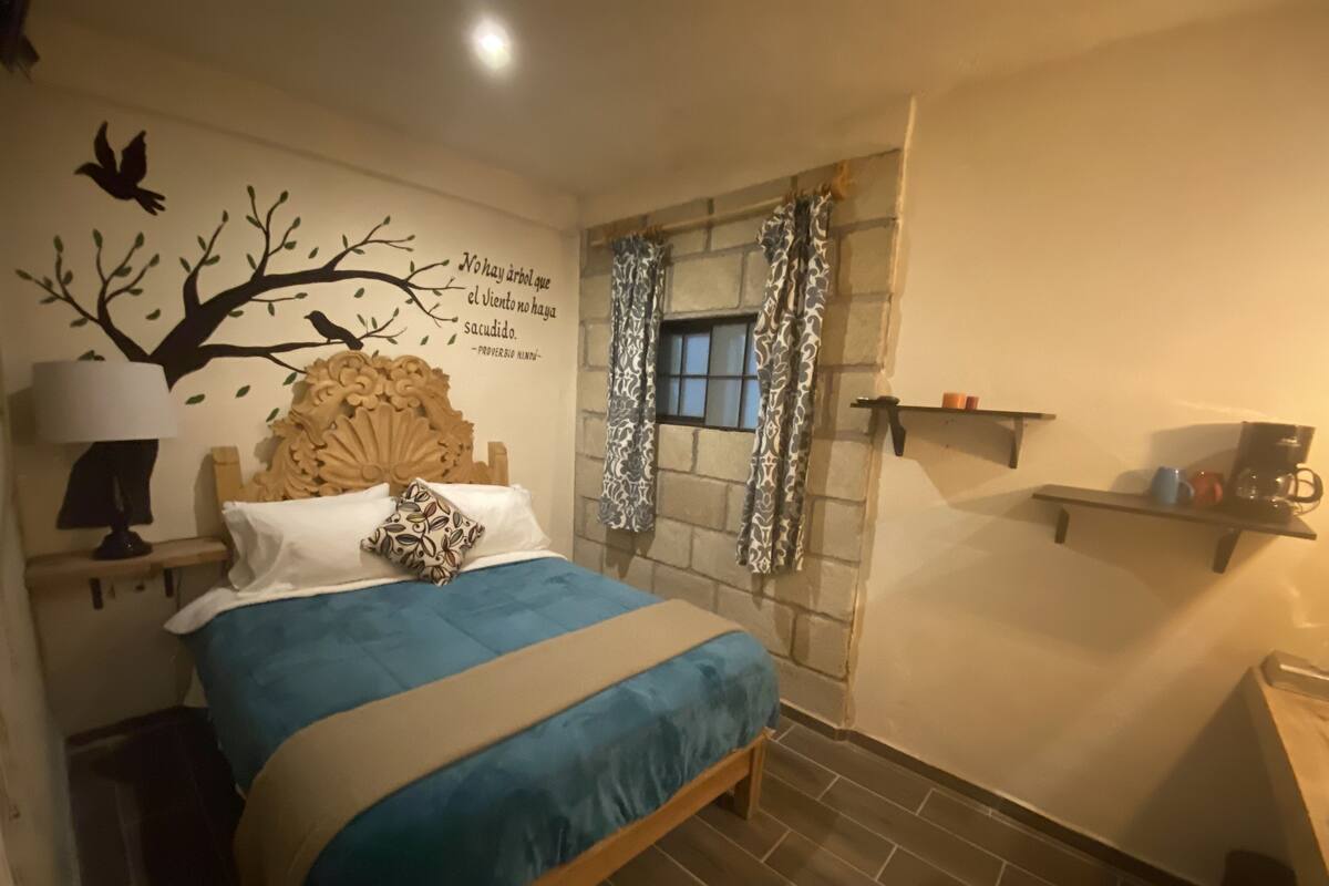 Ein Schlafzimmer mit einem Bett, einer Wandlampe, einer dekorativen Baum-Abbildung, einem Fenster mit gemusterten Vorhängen und einem Regal mit Gegenständen.