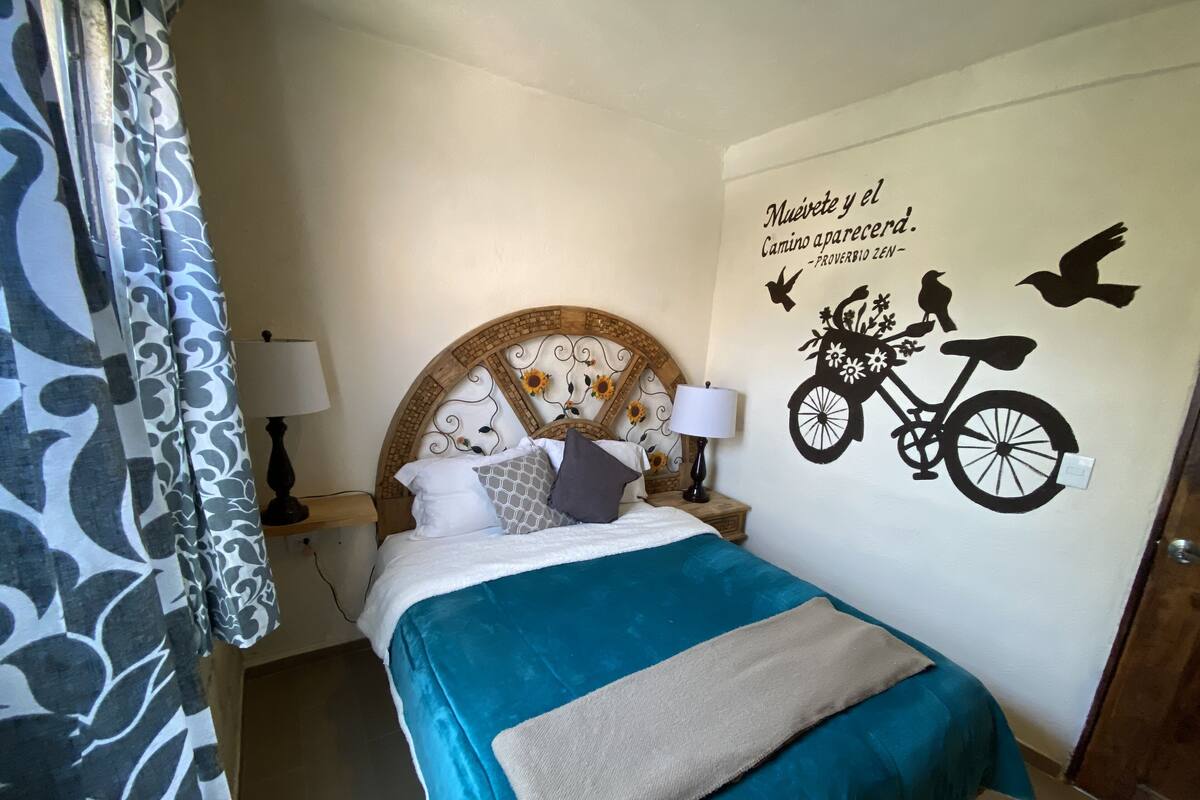 Ein ordentlich bezogenes Bett mit blauer Bettdecke und beiger Tagesdecke, ein Holz-Kopfteil mit floralen Mustern, eine Wandsticker-Darstellung von einem Fahrrad und Vögeln sowie ein gemustertes Vorhang.